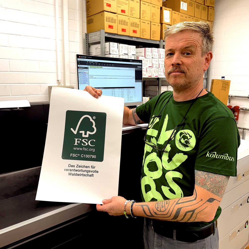 ein freundlicher mann in einem grünen kolumbus t-shirt hält ein blatt mit dem fsc-label hoch